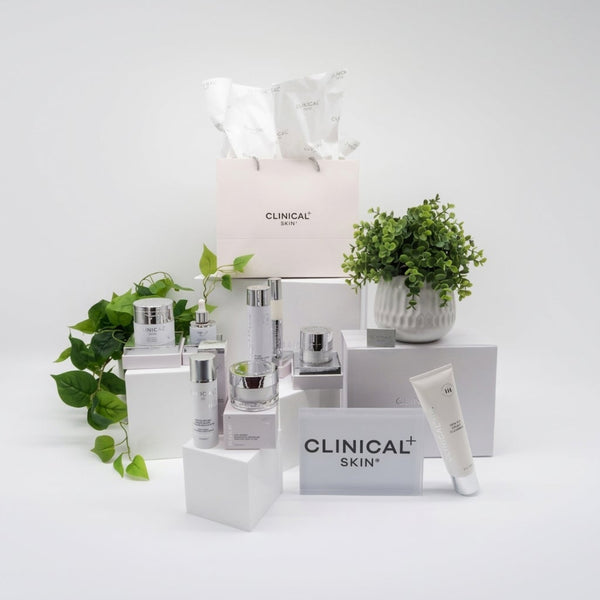 Clinical Skin Soft Skin Reset Routine – complete hydrating skincare set with Clinical Skin moisturizers, serums, and cleanser arranged on white platforms with branded shopping bag and green plants on gray background