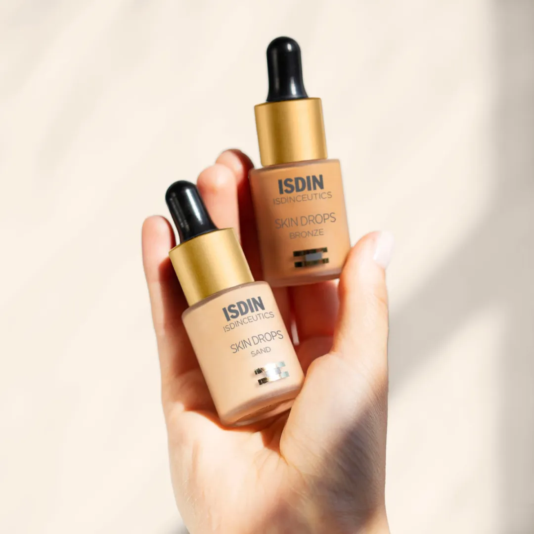 Hand holding ISDIN Skin Drops Bronze bottle beside Skin Drops Sand bottle