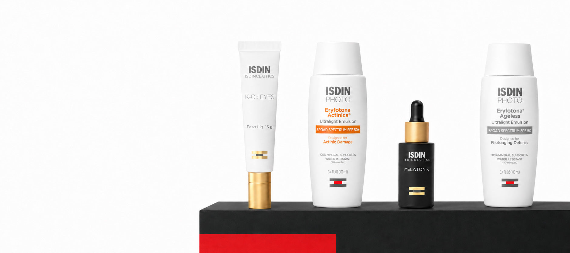 Four ISDIN skincare products arranged on a black platform with a red base against a white background: a white tube with a gold cap, a white bottle with orange accents, a small black dropper bottle with a gold collar, and a large white bottle.