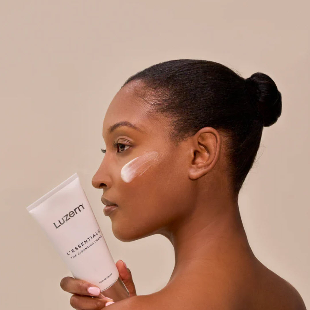 Woman holding a tube of Luzern L'Essentials Pure Cleansing Creme near her cheek with product applied