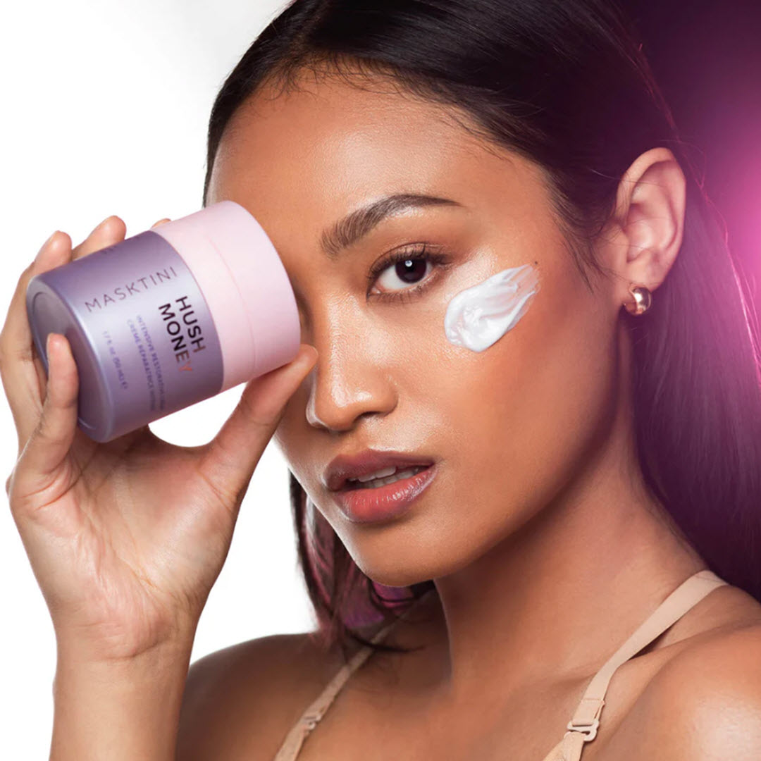 Woman holding a jar of Masktini Hush Money Intensive Restorative Crème with a swatch of the cream under her eye