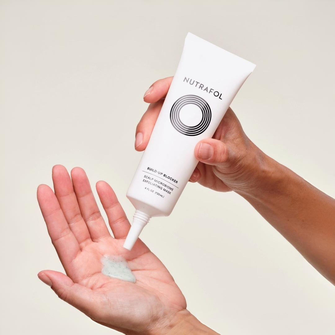 Hands squeezing Nutrafol Build-Up Blocker Exfoliating Mask tube dispensing creamy formula onto palm