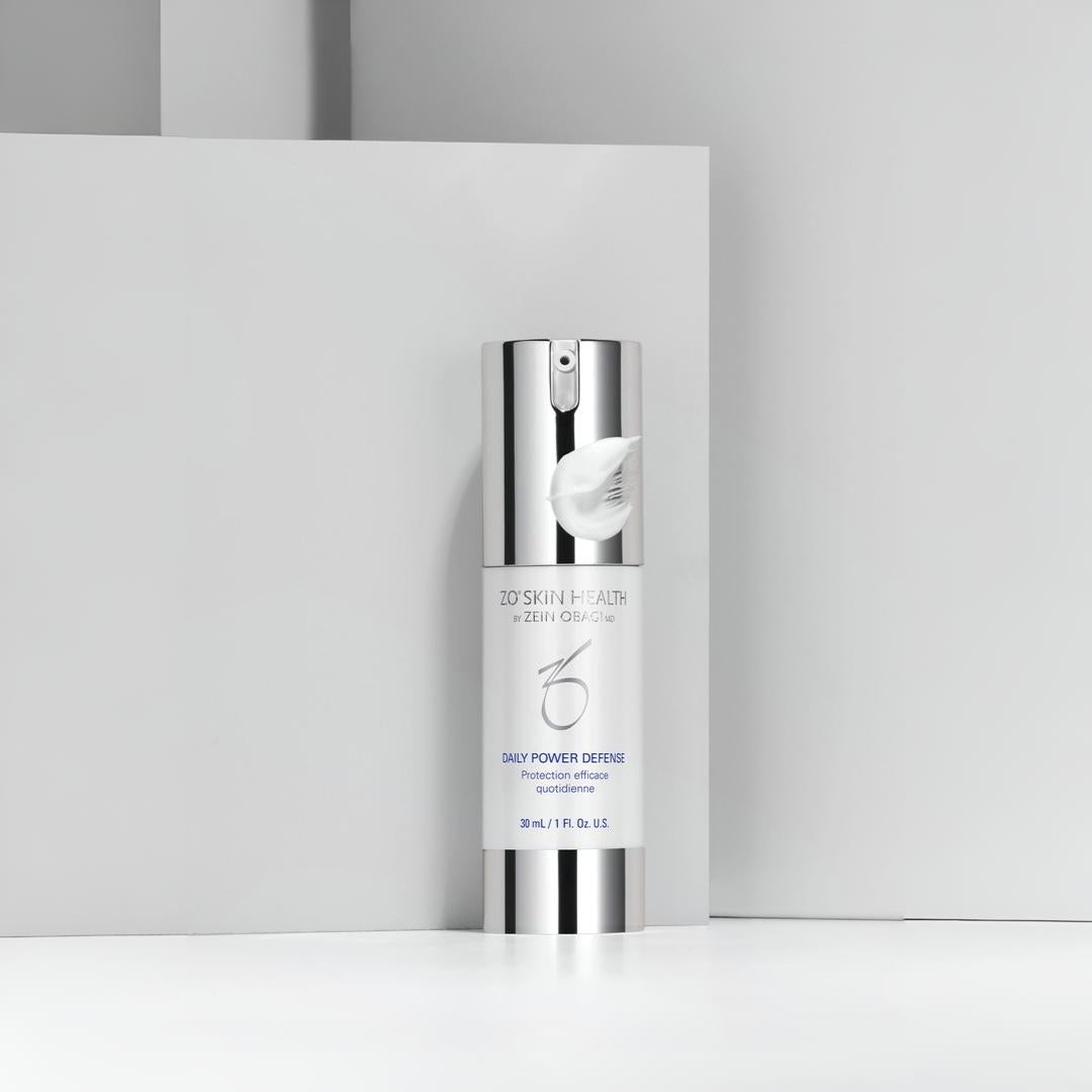 ZO Skin Health Daily Power Defense silver pump bottle with cream swatch against gray geometric backdrop – daily antioxidant protection serum