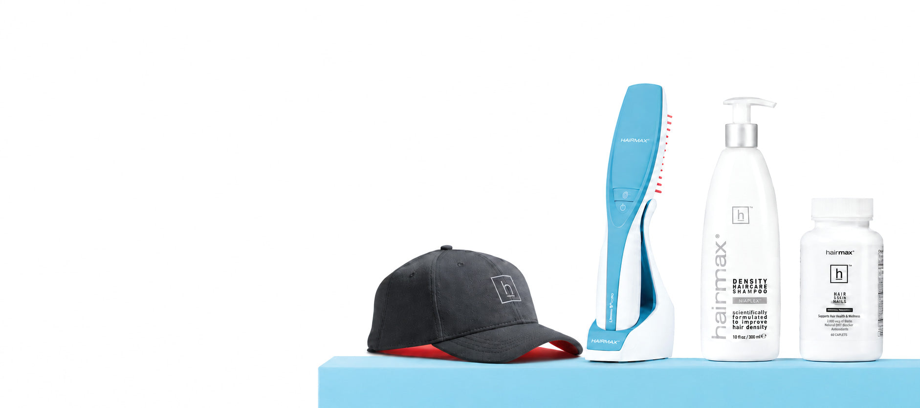 A haircare product lineup on a light blue platform against a white background, featuring a black and red baseball cap, a blue and white hair tool, a white pump bottle labeled hairmax, and a white supplement bottle.