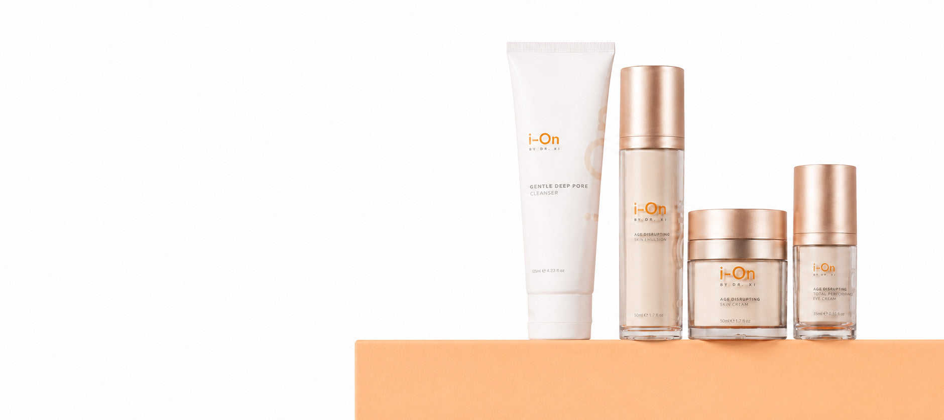 Four skincare products from i-On Beauty arranged on a peach-colored platform against a white background: a white tube, a clear bottle with a rose-gold cap, a clear jar with a rose-gold lid and cream-colored contents, and a small clear bottle with a rose-gold cap.
