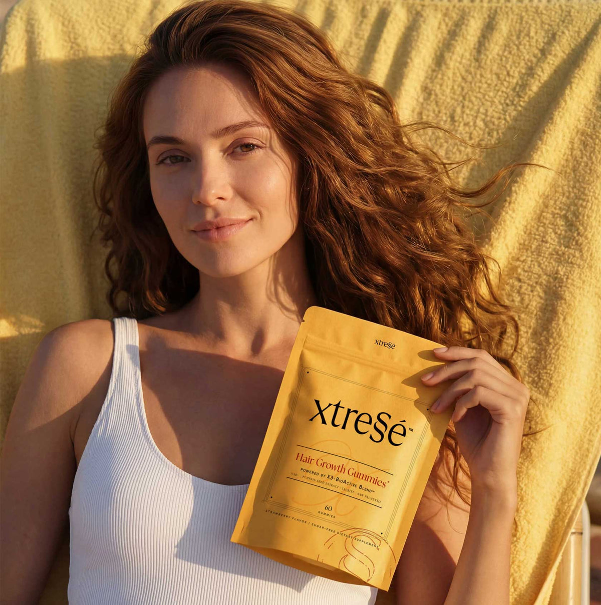 Advanced Dermatology Xtressé Hair Growth Gummies lifestyle – woman with thick long red wavy hair holding gold gummies pouch in sunlight showing results of dermatologist-developed hair growth supplement